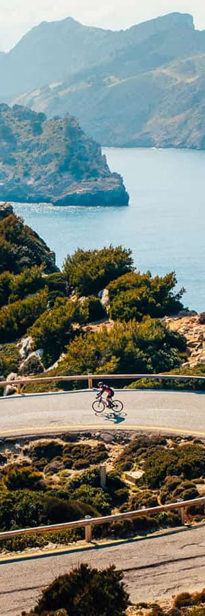Ciclista en carretera costera con monta&ntilde;as y el mar de fondo.